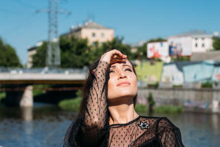 beautiful girl, brunette, in the summer by the river in the city, in a black dress. standing alone...