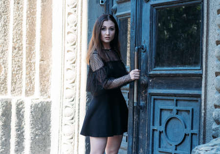 portrait of a beautiful girl, a brunette girl in a black dress, stands near a big black door, goes into it. a new stage in life, enterの写真素材