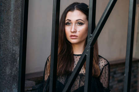 portrait of a young, beautiful girl. The brunette in a black dress is standing near the cage with...