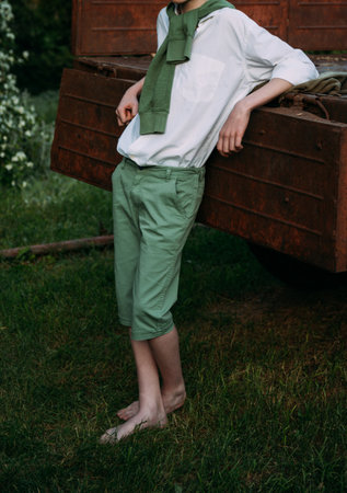 a boy in a white shirt and green pants, a green sweater around his neck, leaning his elbows on the old truck cousin, barefoot on the grass in the summer. village, farmer, on farmの写真素材