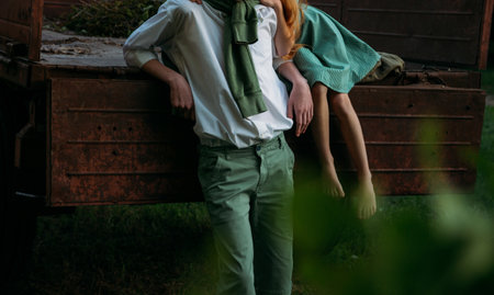 a barefooted girl in a green, vintage dress, the dress sits on the back of a red, old car and hugs the barefoot neck, next to a standing guy who is dressed in a white shirt, green shorts with a green sweater tied around his neck. summer day in the village. teenagers in the village. first love and romance on the grass. rural life of adolescents. purity and innocence, farmerの写真素材