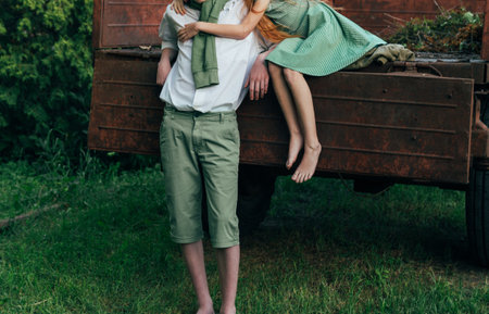 a barefooted girl in a green, vintage dress, the dress sits on the back of a red, old car and hugs the barefoot neck, next to a standing guy who is dressed in a white shirt, green shorts with a green sweater tied around his neck. summer day in the village. teenagers in the village. first love and romance on the grass. rural life of adolescents. purity and innocence, farmerの写真素材