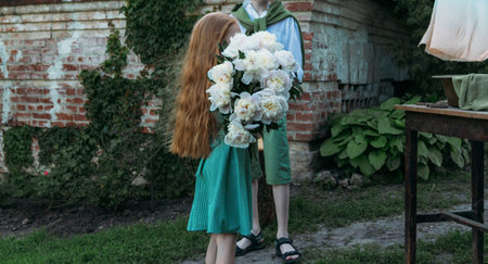 The redhead girl goes with a big bouquet of summer flowers, of white flowers, peonies. girl in vintage green dress. next to it there is a boy in a white shirt and green shorts, with a green sweater. congratulation. give birthday presentsの写真素材