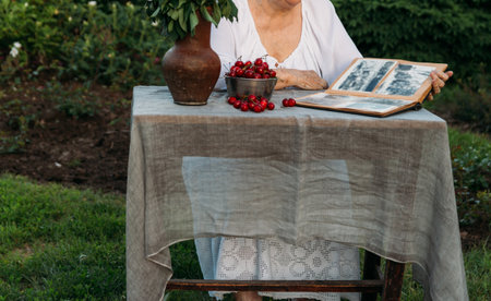grandmother sitting in a chair at the table on which the cherry is lying, in the garden and watching an old album with photos, recounts and remembers the family's story. grandmother in a white vintage dress, summer, warm day. view album with photosの写真素材
