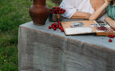 grandmother with her granddaughter sitting in a chair at the table on which the cherry is lying, in the garden and watching an old album with photos, recounts and remembers the family's story. grandmother in a white vintage dress, granddaughter in a green dress summer, warm day. view album with photosの写真素材