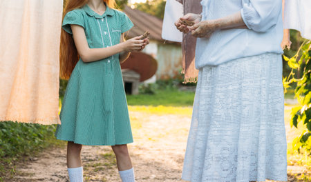 girl next to an elderly woman. granddaughter helps her grandmother. a girl in a green dress is holding clothespins for wet clothes. next to her is a grandmother in white vintage clothing and also holds clothespins, they are about to hang clothes on the rope drying. village and summer. help the elders and relatives. to visit grandmother in the village.の写真素材