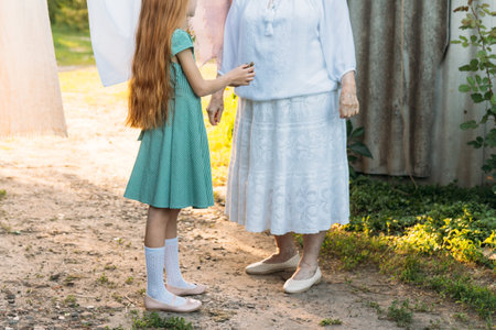 girl next to an elderly woman. granddaughter helps her grandmother. a girl in a green dress is holding clothespins for wet clothes. next to her is a grandmother in white vintage clothing and also holds clothespins, they are about to hang clothes on the rope drying. village and summer. help the elders and relatives. to visit grandmother in the village.の写真素材