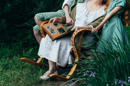 grandmother with her grandchildren sitting in a rocking chair in the garden and watching an old album with photos, recounts and remembers the family's story. grandmother with her grandchildren sitting in a rocking chair in the garden and watching an old album with photos, recounts and remembers the family's story. grandmother in a white vintage dress, granddaughter with tearful hair in a green dress, and a boy in green shorts, a white shirt and a green sweater. summer, warm day. view album with photosの写真素材