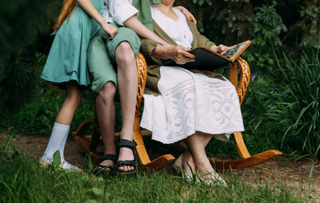 grandmother with her grandchildren sitting in a rocking chair in the garden and watching an old album with photos, recounts and remembers the family's story. grandmother with her grandchildren sitting in a rocking chair in the garden and watching an old album with photos, recounts and remembers the family's story. grandmother in a white vintage dress, granddaughter with tearful hair in a green dress, and a boy in green shorts, a white shirt and a green sweater. summer, warm day. view album with photosの写真素材