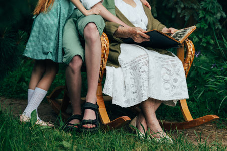 grandmother with her grandchildren sitting in a rocking chair in the garden and watching an old album with photos, recounts and remembers the family's story. grandmother with her grandchildren sitting in a rocking chair in the garden and watching an old album with photos, recounts and remembers the family's story. grandmother in a white vintage dress, granddaughter with tearful hair in a green dress, and a boy in green shorts, a white shirt and a green sweater. summer, warm day. view album with photosの写真素材