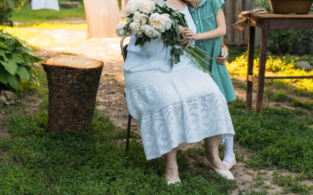 grandmother, an elderly woman in white, vintage clothes sitting and hugging her granddaughter, a girl in a green dress, and holding in her other hand a bouquet of flowers, white peonies, in the garden. meeting and congratulations, a giftの写真素材