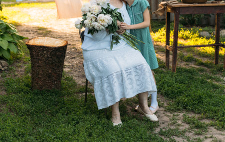 grandmother, an elderly woman in white, vintage clothes sitting and hugging her granddaughter, a girl in a green dress, and holding in her other hand a bouquet of flowers, white peonies, in the garden. meeting and congratulations, a giftの写真素材
