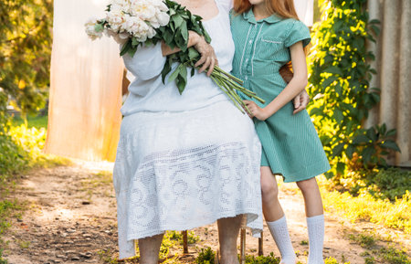 respect of elders. meeting of relatives. grandmother, an elderly woman in white, vintage clothes sitting and hugging her granddaughter, a girl in a green dress, and holding in her other hand a bouquet of flowers, white peonies, in the garden. meeting and congratulations, a giftの写真素材