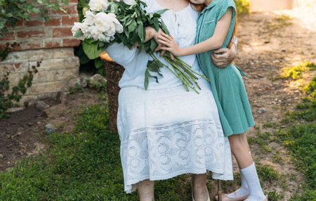 family ties. grandmother, an elderly woman in white, vintage clothes sitting and hugging her granddaughter, a girl in a green dress, and holding in her other hand a bouquet of flowers, white peonies, in the garden. meeting and congratulations, a giftの写真素材
