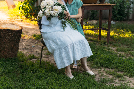 visit grandmother. grandmother, an elderly woman in white, vintage clothes sitting and hugging her granddaughter, a girl in a green dress, and holding in her other hand a bouquet of flowers, white peonies, in the garden. meeting and congratulations, a giftの写真素材