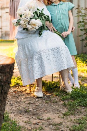 love between grandmother and granddaughter. grandmother, an elderly woman in white, vintage clothes sitting and hugging her granddaughter, a girl in a green dress, and holding in her other hand a bouquet of flowers, white peonies, in the garden. meeting and congratulations, a giftの写真素材
