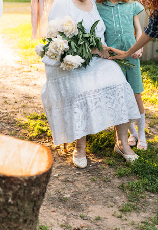 grandmother, an elderly woman in white, vintage clothes sitting and hugging her granddaughter, a girl in a green dress, and holding in her other hand a bouquet of flowers, white peonies, in the garden. meeting and congratulations, a gift.a female hand reaches for a bouquet of flowersの写真素材