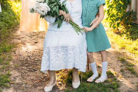 grandmother, an elderly woman in white, vintage clothes sitting and hugging her granddaughter, a girl in a green dress, and holding in her other hand a bouquet of flowers, white peonies, in the garden. meeting and congratulations, a giftの写真素材