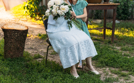 grandmother, an elderly woman in white, vintage clothes sitting and hugging her granddaughter, a girl in a green dress, and holding in her other hand a bouquet of flowers, white peonies, in the garden. meeting and congratulations, a giftの写真素材