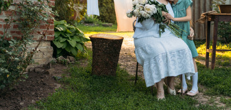 grandmother, an elderly woman in white, vintage clothes sitting and hugging her granddaughter, a girl in a green dress, and holding in her other hand a bouquet of flowers, white peonies, in the garden. meeting and congratulations, a giftの写真素材