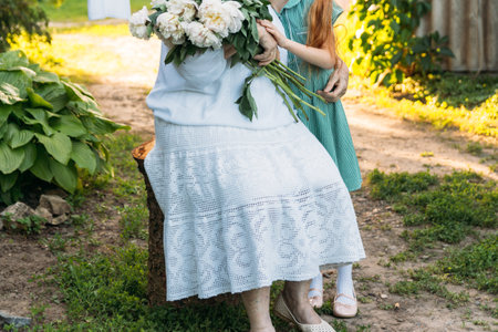 grandmother, an elderly woman in white, vintage clothes sitting and hugging her granddaughter, a girl in a green dress, and holding in her other hand a bouquet of flowers, white peonies, in the garden. meeting and congratulations, a giftの写真素材