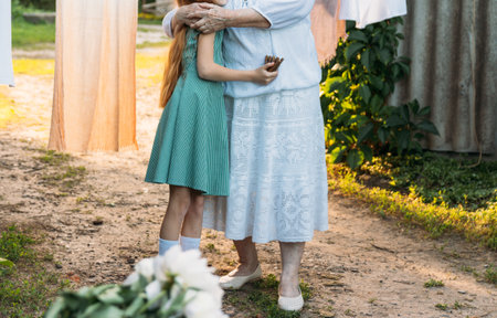 an elderly woman in white, vintage clothes stands and hugs her granddaughter, a girl in a green dress, in the garden. meeting and congratulations, a gift, on the stump is a bouquet of white peonies. in the backgroundの写真素材