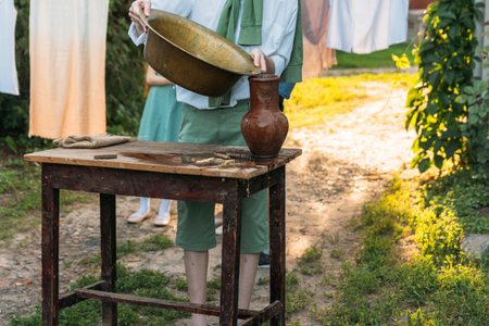 a boy in a white shirt and a green sweater, holds a clay jug, pours water out of it into a copper basin. help, countryside. village.の写真素材