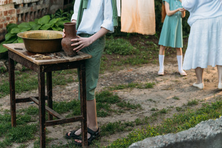 a boy in a white shirt and a green sweater, holds a clay jug, pours water out of it into a copper basin. help, countryside. village. in the background is an elderly woman and a girl, hang clothesの写真素材
