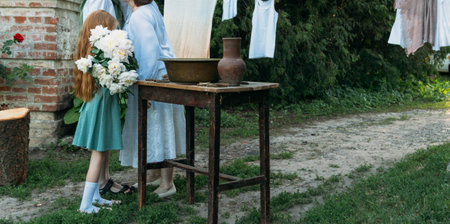 meeting grandmother and grandchildren. grandmother embraces grandchildren, brother and sister. grandson and granddaughter came to visit. give flowers. relatives meeting. Grandmother in white vintage clothing and grandchildren in green clothes. next to it there is a table on which lie clothes for washing by handsの写真素材