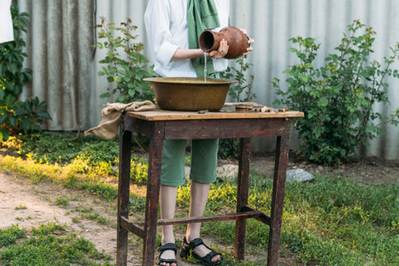 a boy in a white shirt and a green sweater, holds a clay jug, pours water out of it into a copper...