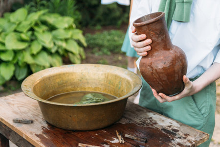 a boy in a white shirt and a green sweater, holds a clay jug, pours water out of it into a copper basin. help, countryside. village.の写真素材