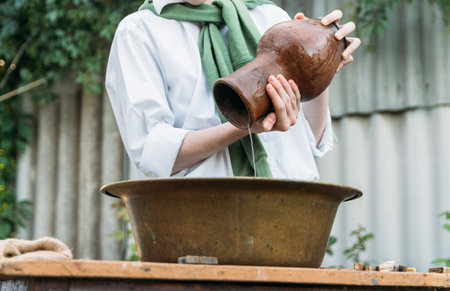 a boy in a white shirt and a green sweater, holds a clay jug, pours water out of it into a copper basin. help, countryside. village.の写真素材