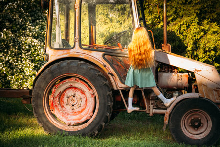 a little girl in a green dress with red hair climbs, climbed up, stands on the cab of a red, old tractor. on a farm in the summer. in the village in the summer. back view. the hair falls on the sunの写真素材