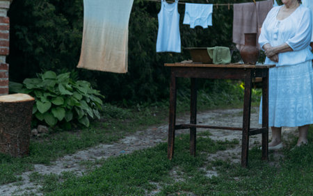 an old woman stands and waits, in white vintage clothes, in the courtyard, near the table. on the table is an old clay jug and an old copper basin with water and cloth. next are the clothespins. Behind hangs, clothes are dried. village wash.の写真素材