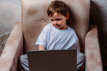 little boy in a white t-shirt sits on a chair, sofa with a laptop. looks into the camera. funny Face. concentration.の写真素材