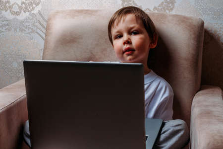 little boy in a white t-shirt sits on a chair, sofa with a laptop. looks into the camera. funny Face. concentration.の写真素材