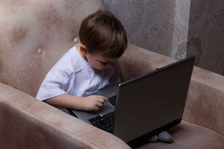 little boy in a white t-shirt sits on a chair, sofa with a laptop. looks into the camera. funny Face. concentration.の写真素材