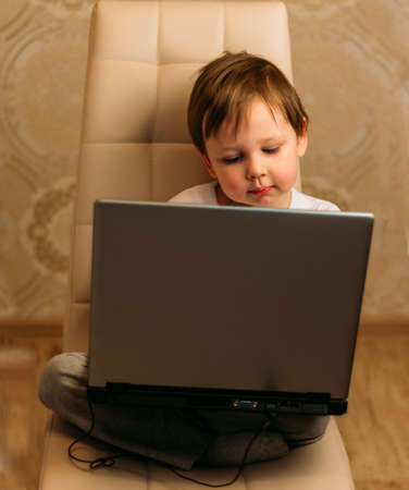 little boy in a white t-shirt sits on a chair, sofa with a laptop. looks into the camera. funny Face. concentration.の写真素材