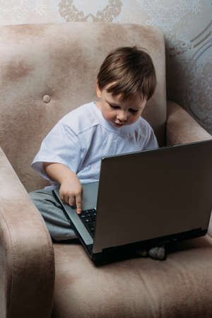 little boy in a white t-shirt sits on a chair, sofa with a laptop. looks into the camera. funny Face. concentration.の写真素材