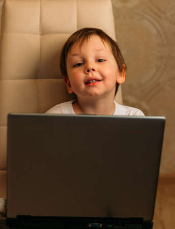 little boy in a white t-shirt sits on a chair, sofa with a laptop. looks into the camera. funny Face. concentration.の写真素材