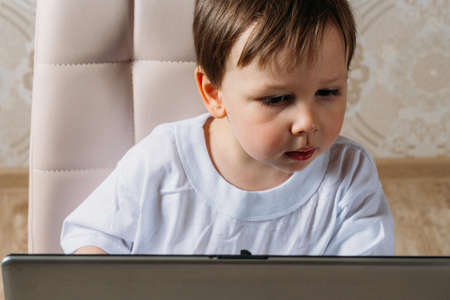 little boy in a white t-shirt sits on a chair, sofa with a laptop. looks into the camera. funny Face. concentration.の写真素材