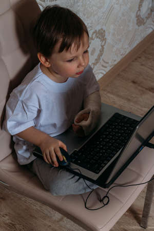 little boy in a white t-shirt sits on a chair, sofa with a laptop. looks into the camera. funny Face. concentration.の写真素材