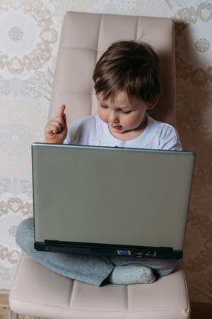 little boy in a white t-shirt sits on a chair, sofa with a laptop. looks into the camera. funny Face. concentration.の写真素材