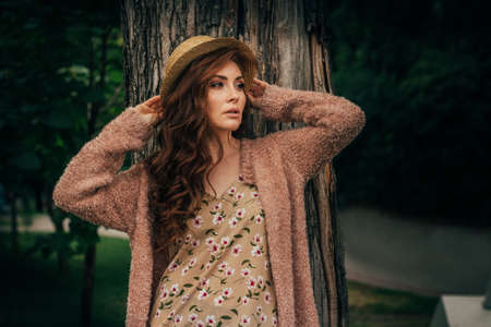portrait of a beautiful girl who stands near a tree. summer, walk in nature, red-haired girl in a vintage dress and a beige sweater.in Hat romance and flirtの写真素材