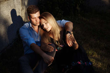 couple in love is sitting on the grass near the wall, the sun is shining in her eyes, her eyes are...