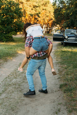 the girl jumped on the guy's back.he holds her legs.check shirt and jeans.in the background is a black car in the forestの写真素材