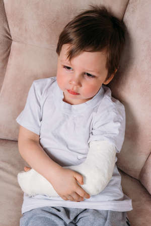 a small boy in a white T-shirt sits on a chair with a broken arm, in a cast. hospital. medicine. danger. pain. to break limbs. violence.の写真素材
