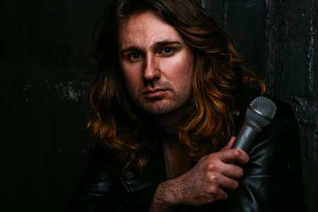 a portrait of a brutal, handsome man, a singer who sits on a chair on a dark background, in a black leather kurt on his naked body and holds a microphone in his hands. presenter, singer at the concert. in blue jeansの写真素材