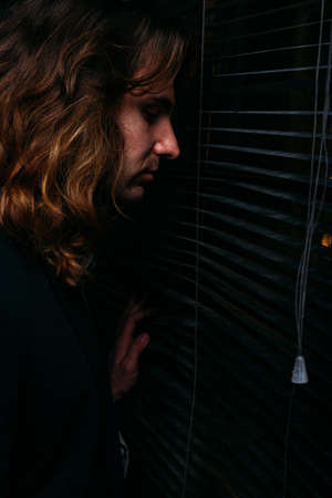 a man with long hair is standing near the window, opening the blinds with lobes, in a black jacket....
