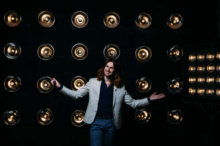 a man in a white jacket and in a black shirt, with long hair, stands against the backdrop of bright...